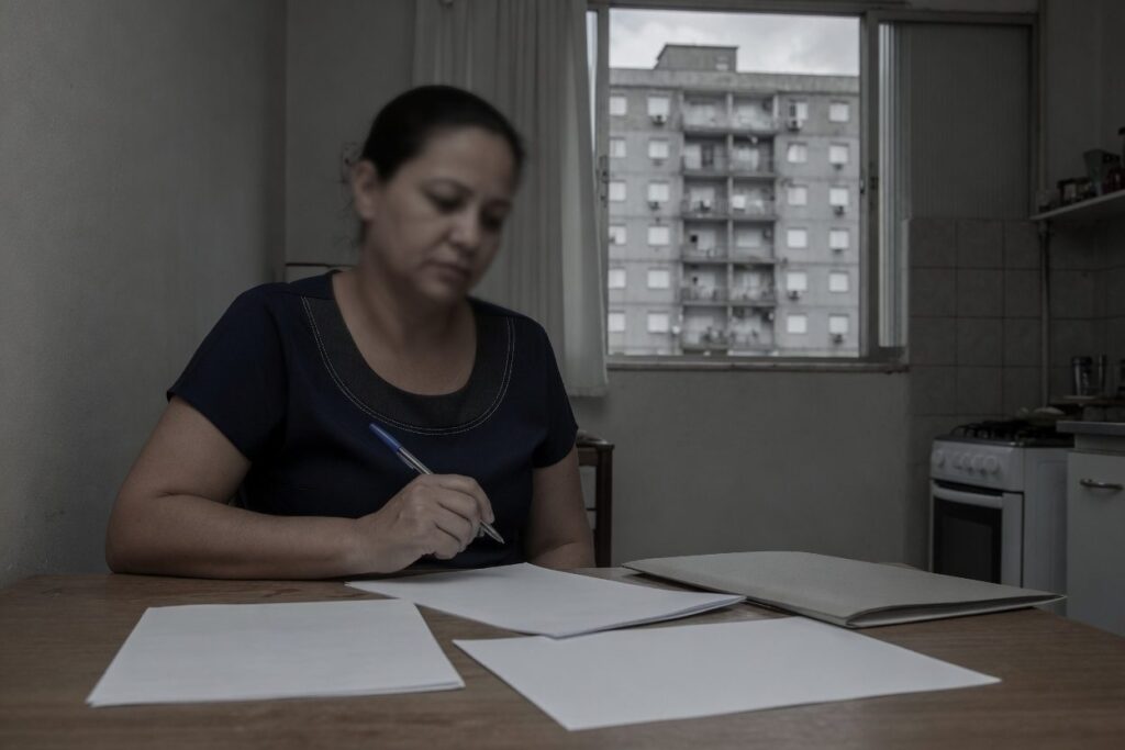 Mulher revisando documentos na cozinha com prédio residencial ao fundo, ilustrando dívidas que acompanham o imóvel