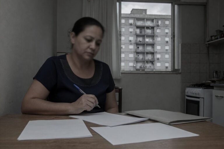 Mulher revisando documentos na cozinha com prédio residencial ao fundo, ilustrando dívidas que acompanham o imóvel